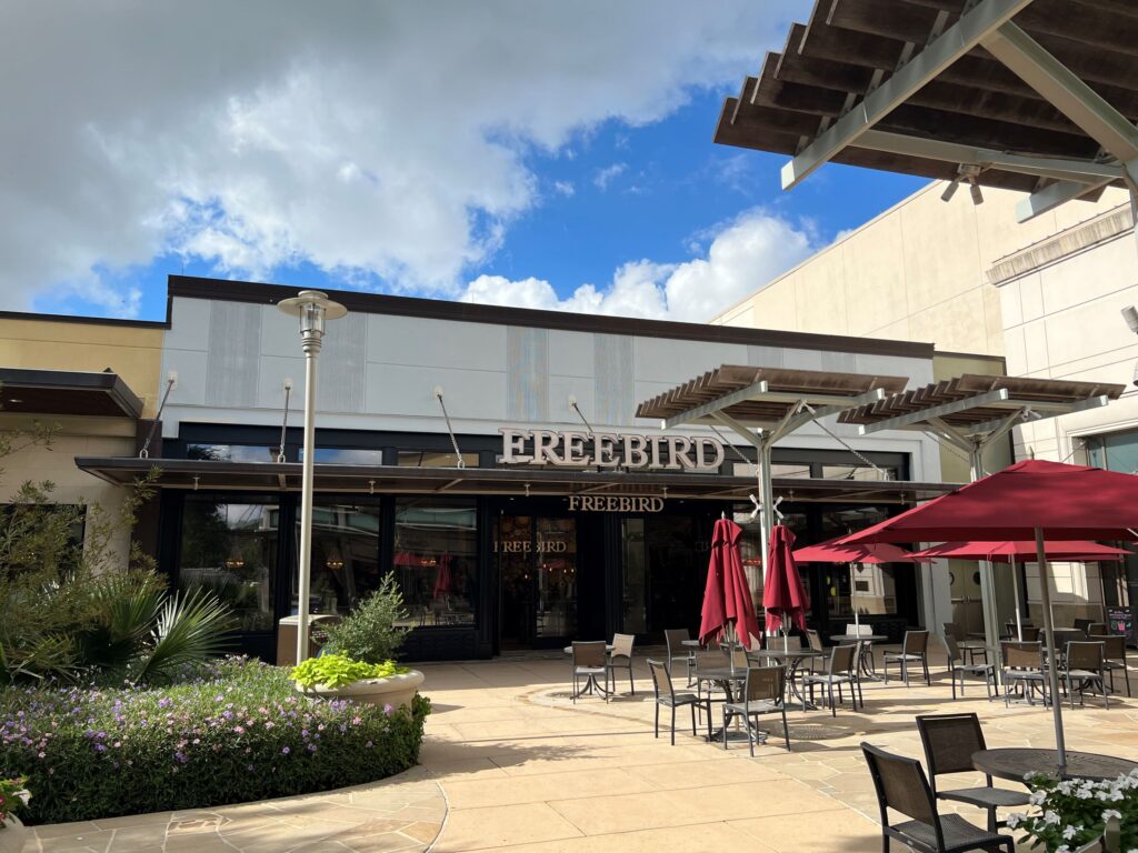 Outdoor seating area with red umbrellas in front of a restaurant named "Freebird" on a sunny day with scattered clouds, constructed by D Barnes Construction.