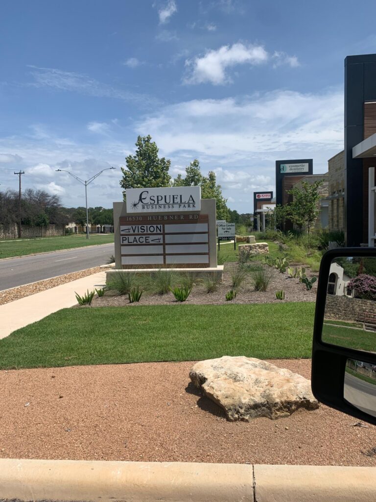 A sunny day view from a vehicle capturing a landscaped area with a sign that reads "Espuela" and "Vision Place" on a roadside, featuring D Barnes Construction buildings in the background.