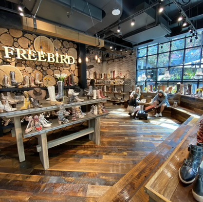 An inviting boutique shoe store with a rustic wooden interior by D Barnes Construction, showcasing a variety of footwear on shelves against a backdrop with a log-filled wall, as customers peruse the selection.